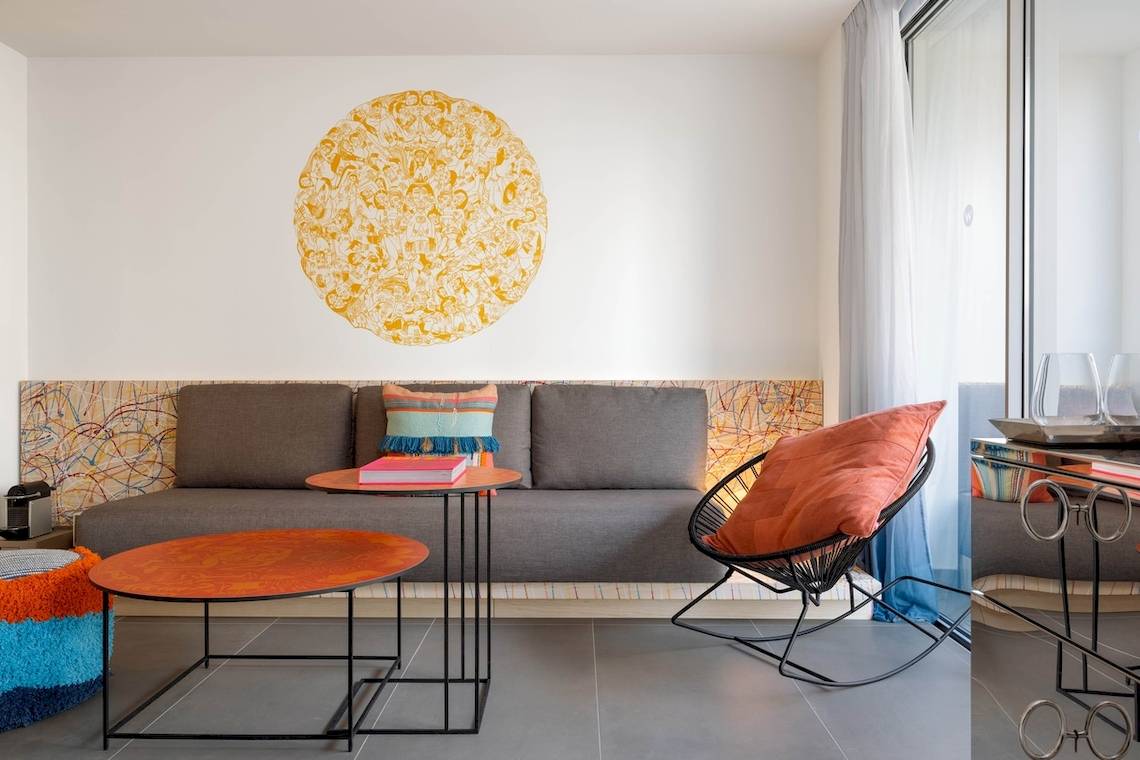 Innenansicht des Wohnzimmers im W Ibiza Hotel mit grauem Sofa, orangefarbenem Stuhl und runden Tischen.