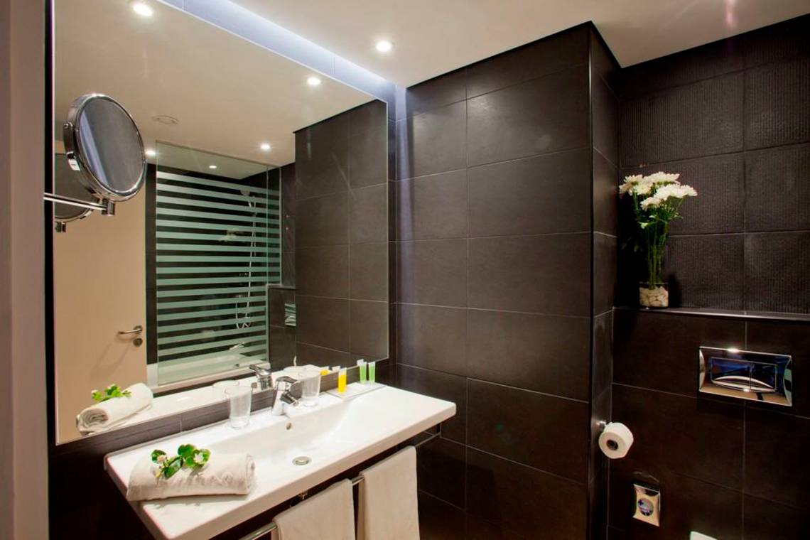 Badezimmer im Faros Hotel Ayia Napa mit schwarzem Wandfliesen, weißem Waschbecken und Spiegel.