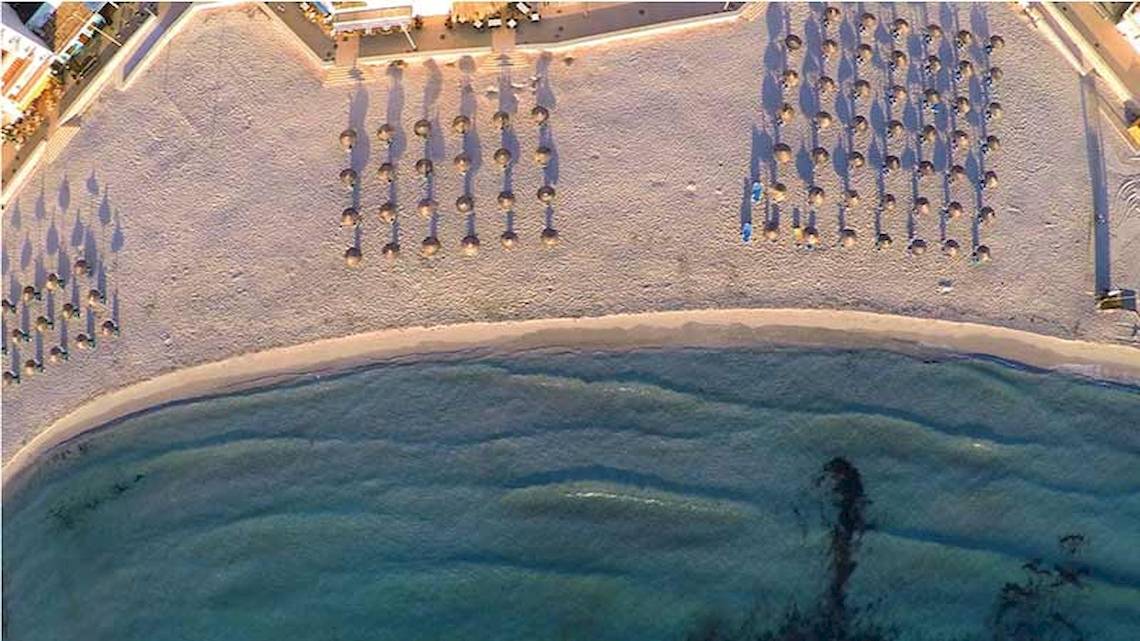 Luftaufnahme der Perla Hotel Strand mit vielen Sonnenschirmen und Gästen am Meer