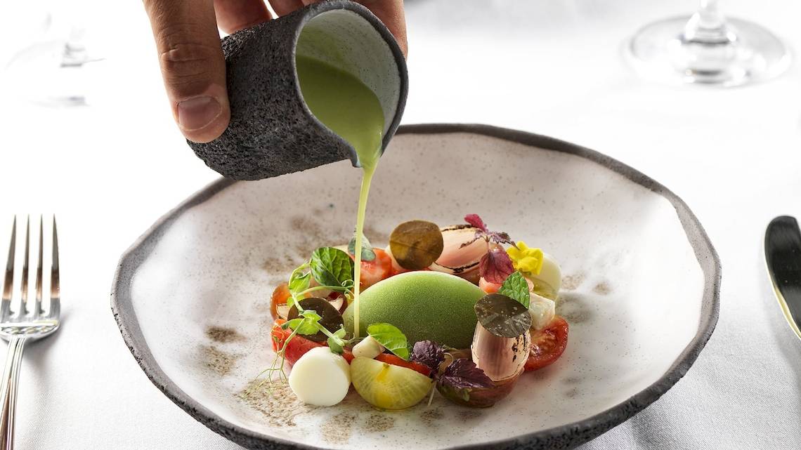 Ein Tellergericht wird mit grüner Flüssigkeit aus einem Becher übergossen, serviert auf einem weißen Tisch.