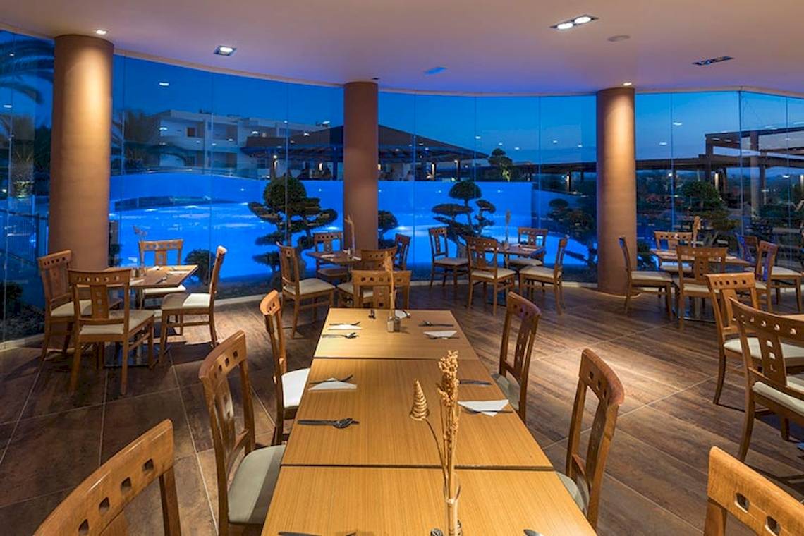 Innenbereich des Sandy Beach Hotel in Kos mit vielen Holztischen und Stühlen und einer großen Glasfassade.