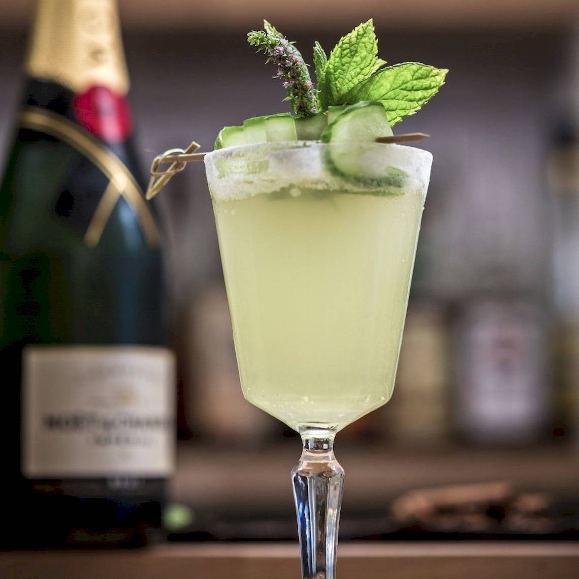 Ein Glas mit einem Cocktail, garniert mit Gurkenscheiben und Minze, vor einer Flasche Champagner.