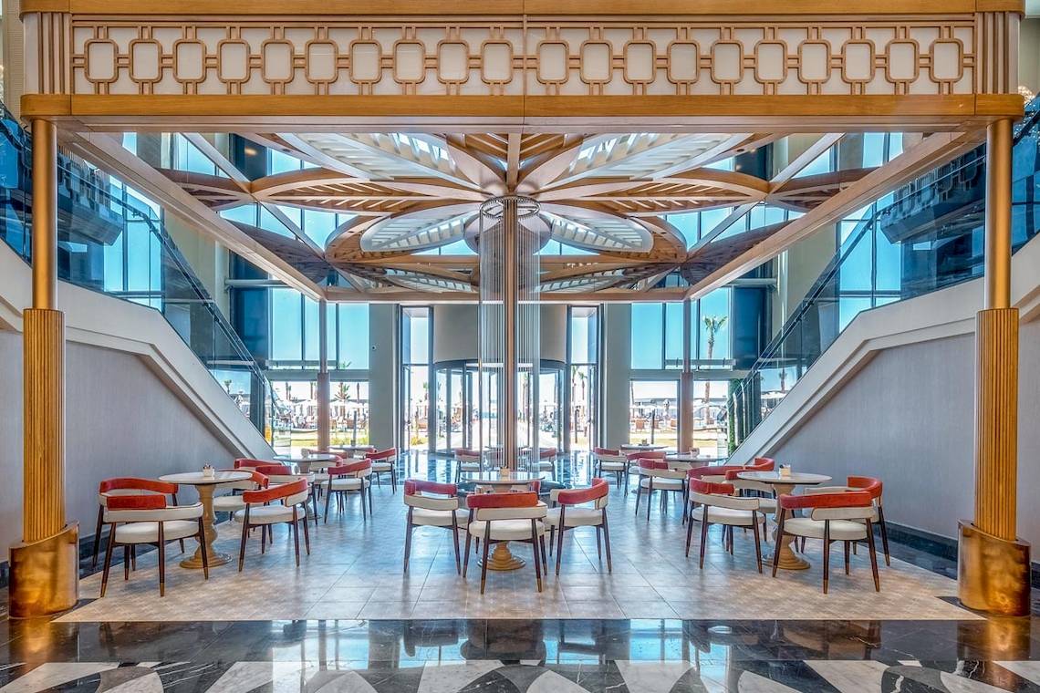 Innenbereich des Lago Hotels mit modernem Design, runden Tischen und Stühlen, Glaswänden und einem zentralen Holzelement.