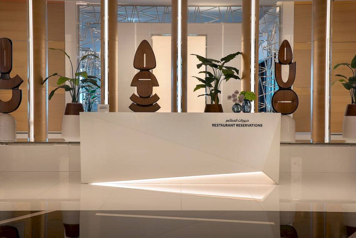 Restaurantreservierungstresen im Marriott Resort Palm Jumeirah, Dubai, mit Pflanzen und modernen Skulpturen im Hintergrund.