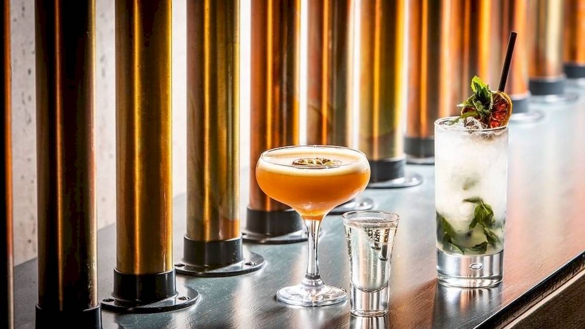 Drei Cocktails auf einer Bar mit goldenen Säulen, darunter ein Orangen-Cocktail, ein Mojito und ein Shot.