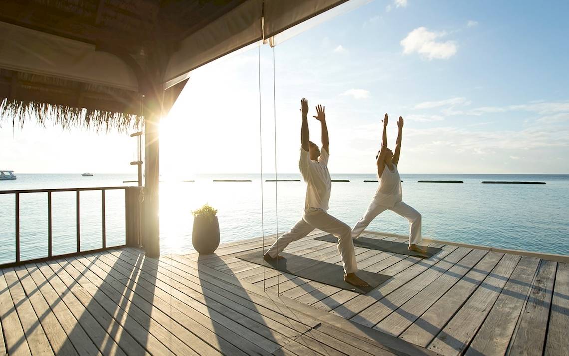 Zwei Personen praktizieren Yoga auf einer Holzplattform vor dem Meer.