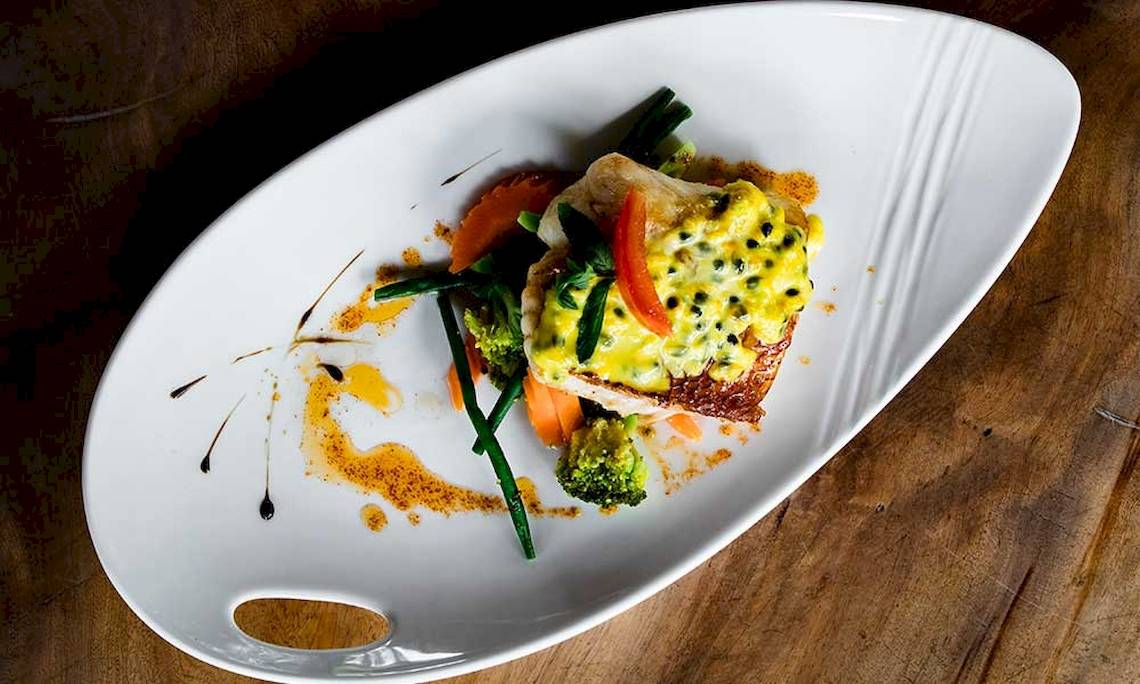 Ein Gericht auf einem weißen Teller auf einem Holztisch, bestehend aus Fisch, Gemüse und Sauce.