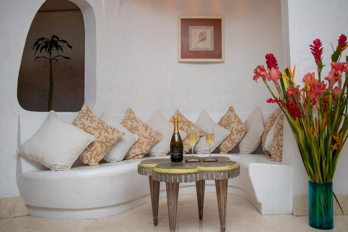 Loungebereich im Village du Pecheur Hotel mit weißer Sitzbank, Kissen, Flasche Champagner und zwei Gläsern auf einem Tisch neben bunten Blumen in einem blauen Vasen.