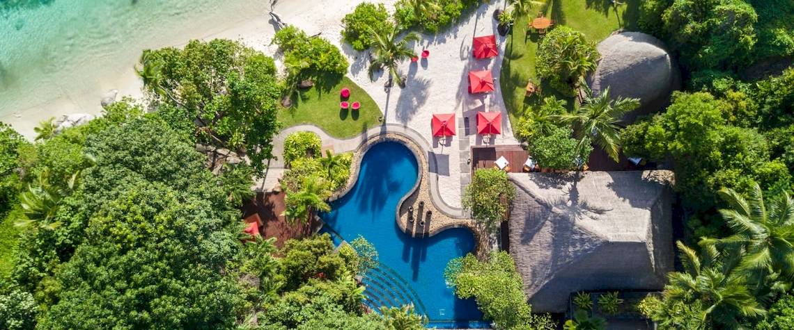 Luftaufnahme des Anantara Maia Seychelles Villas mit Pool, roten Sonnenschirmen und Strand
