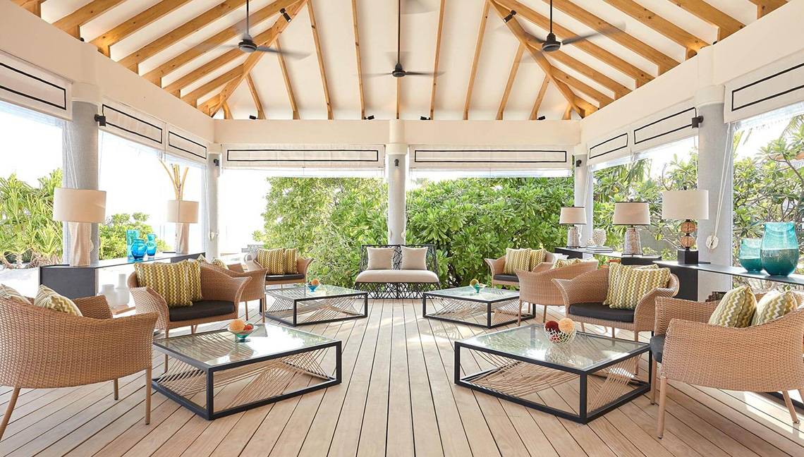 Lounge-Bereich des Amari Havodda Maldives mit Holzboden, modernen Möbeln und großen Fenstern, die einen Blick auf die Außenlandschaft gewähren.