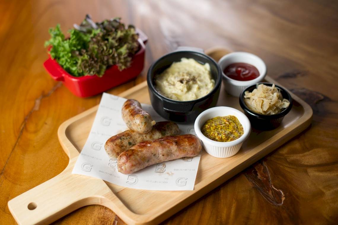 Ein Gericht auf einer Holzplatte im Novotel Bangkok Sukhumvit 20, bestehend aus Würstchen, Kartoffelpüree und verschiedenen Dips.
