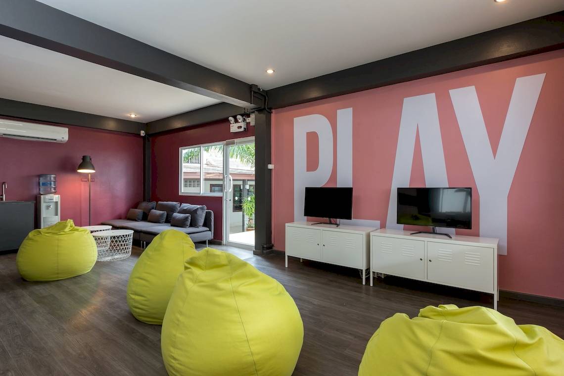 Hotelzimmer mit rosa Wand, gelben Sitzsäcken und Fernsehern, das Wort PLAY auf der Wand.