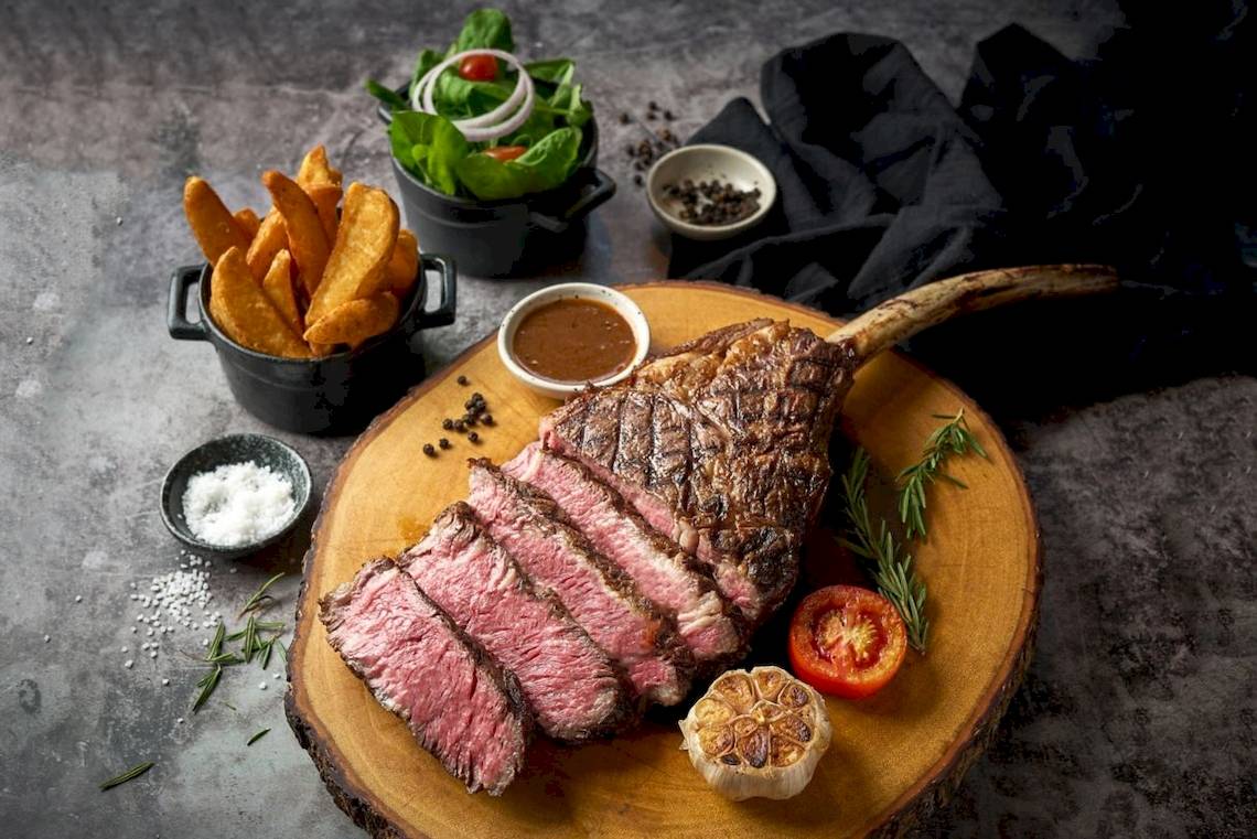 Gegrilltes Steak auf einem Holzbrett serviert mit Beilagen wie Pommes, Salat, Tomaten, Knoblauch und Rosmarin.