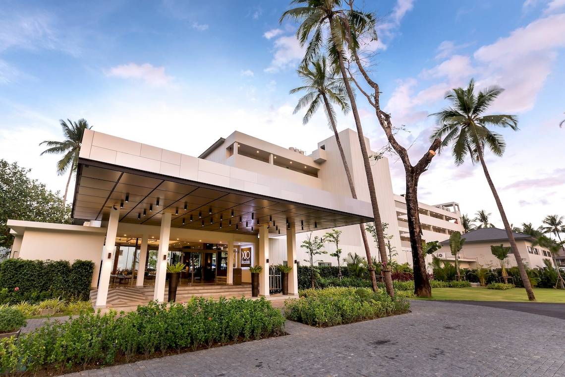 Außenansicht des X10 Khaolak Resorts mit Eingang, Palmen und Sonnenlicht.