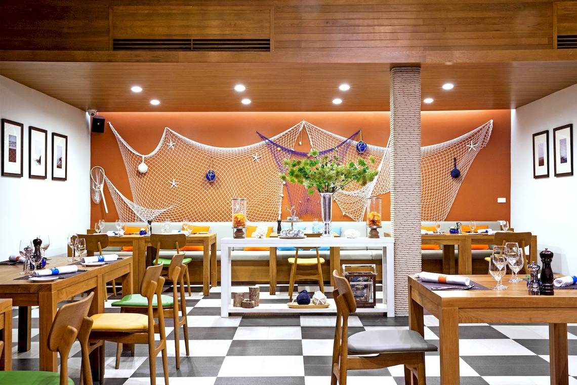 Innenraum des X10 Khaolak Resorts mit oranger Wand, weißen Netzen, schwarzen und weißen Schachbrettboden und mehreren Holztischen.