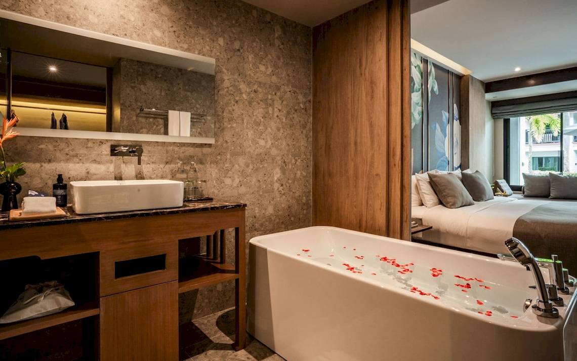 Badezimmer im La Flora Khao Lak Hotel mit Badewanne, Waschbecken und Bett im Hintergrund.
