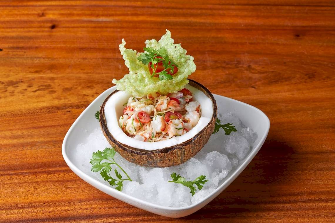 Ein Salat aus Garnelen in einer Kokosnussschale, auf Eis und mit Kräutern garniert, serviert auf einem weißen Teller.