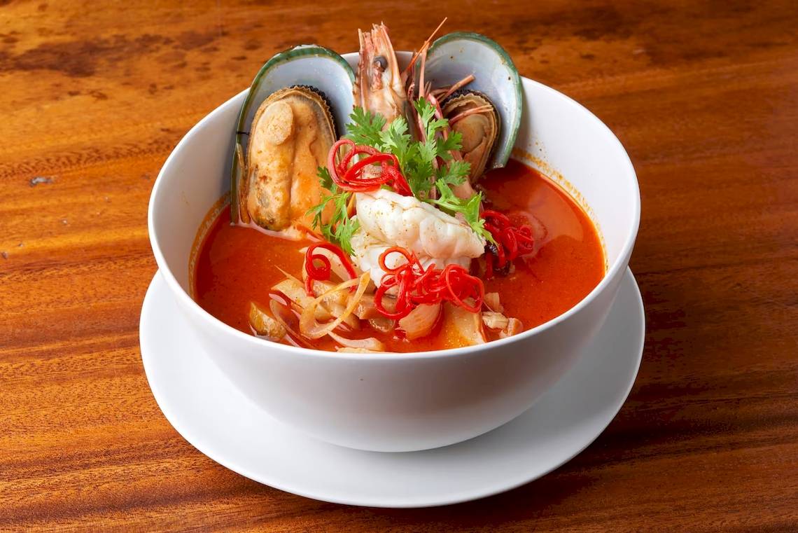 Ein weißer Porzellanschale mit Thai-Suppe, die mit Garnelen, Muscheln und roten Chilis garniert ist.