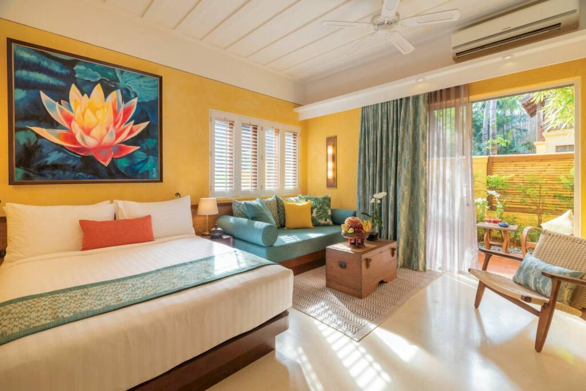 Innenansicht eines Zimmers im Buri Rasa Village Samui Hotel mit Bett, Couch, Kunstwerk und Terrassentür.