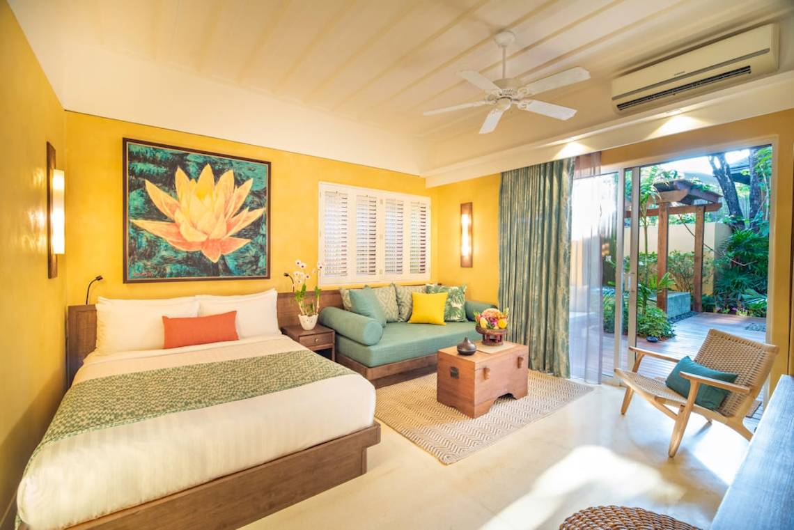 Ein hellgelbes Zimmer im Buri Rasa Village Samui Hotel mit einem Bett, Sofa, Tisch und Gartenblick.