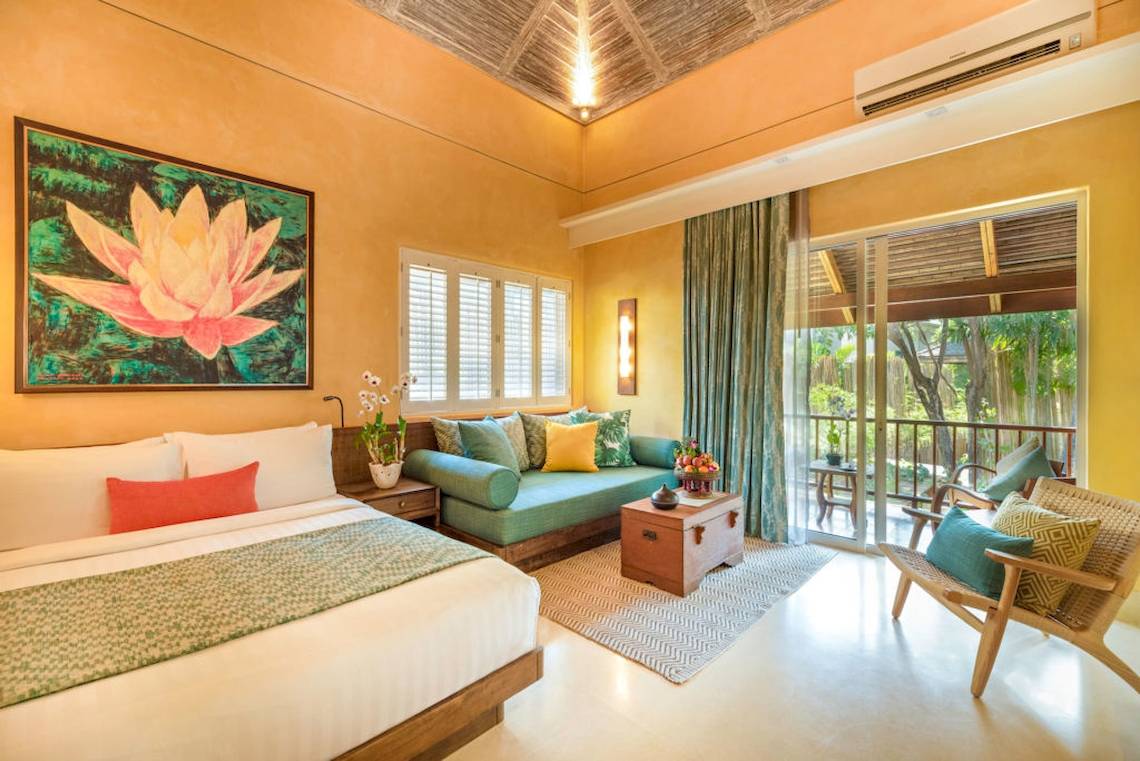 Innenansicht eines Hotelzimmers im Buri Rasa Village Samui, mit einem großen Bett, einem grünen Sofa, einem Tisch und einer Aussicht auf den Garten.