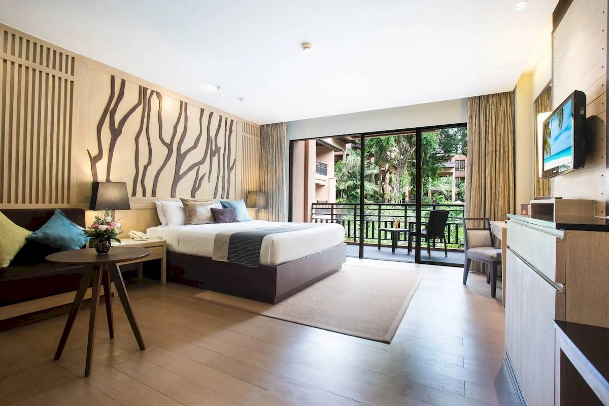 Innenansicht eines modernen Hotelzimmers mit Bett, Couch, Tisch, Fernseher und Balkon.