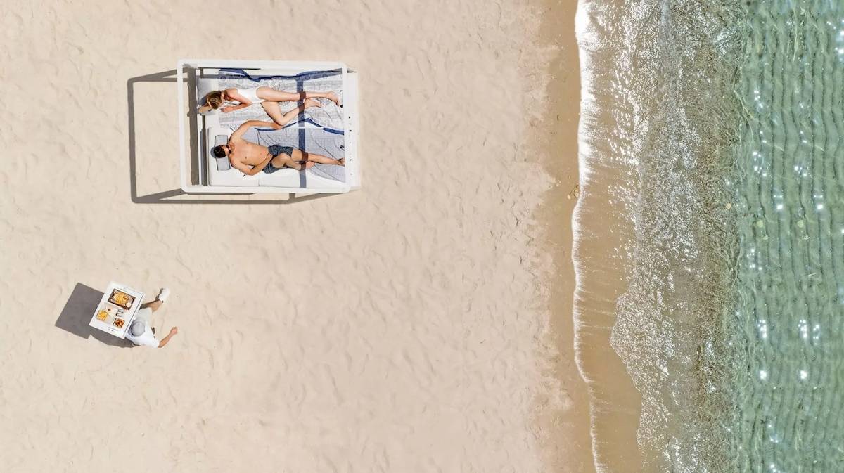 Zwei Personen liegen auf einem Bett am Strand mit einem Servicemitarbeiter, der ein Tablet hält.