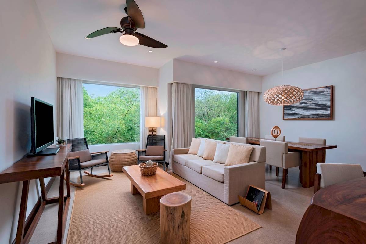 Wohnzimmer mit Fernseher, Sofa, Tisch, Stühlen und großen Fenstern, die auf Bäume hinausgehen.