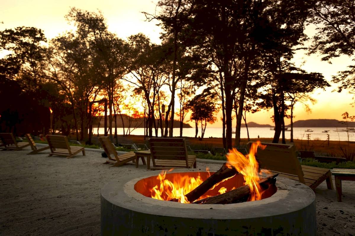 Feuerstelle bei Sonnenuntergang im Freien mit Holzbänken und Blick auf den See im El Mangroove, Autograph Collection Hotel.