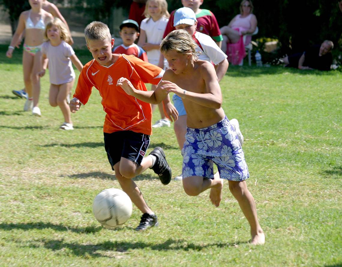 Kinder spielen Fussball auf einer Rasenflaeche, einer juengt verfolgt den Ball.