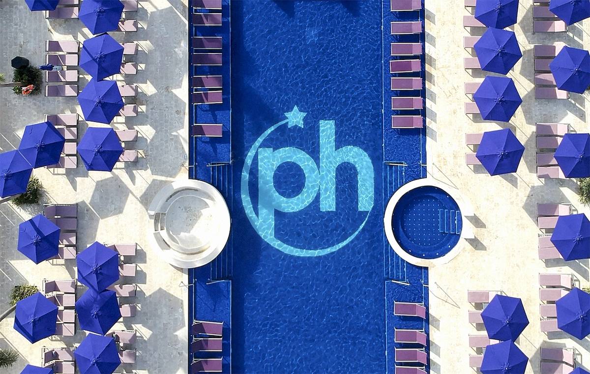 Luftaufnahme des Pools mit blauen Sonnenschirmen und Liegen um den Pool herum, Logo PH im Pool.