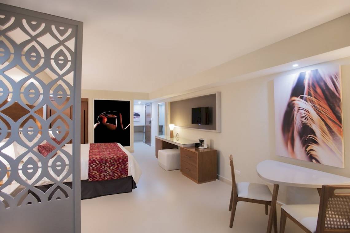Innenansicht eines Hotelzimmers im Emotions By Hodelpa Puerto Plata, mit Bett, Schreibtisch und Kunstwerk.