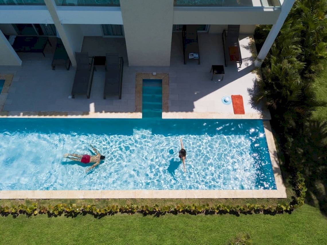 Luftaufnahme von zwei Personen, die im Pool schwimmen, vor einem Hotel mit Terrasse und Liegestühlen.