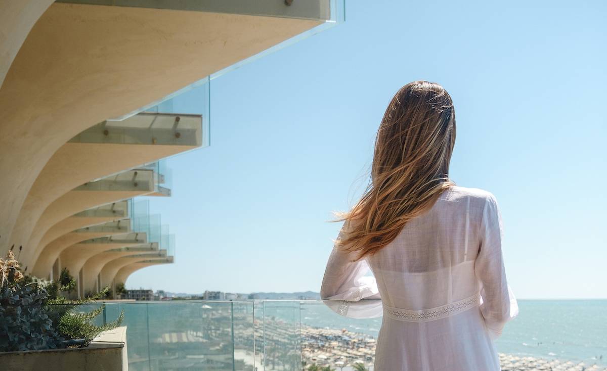 Rückansicht einer Frau in einem weißen Kleid, das auf einem Balkon mit Meerblick steht.