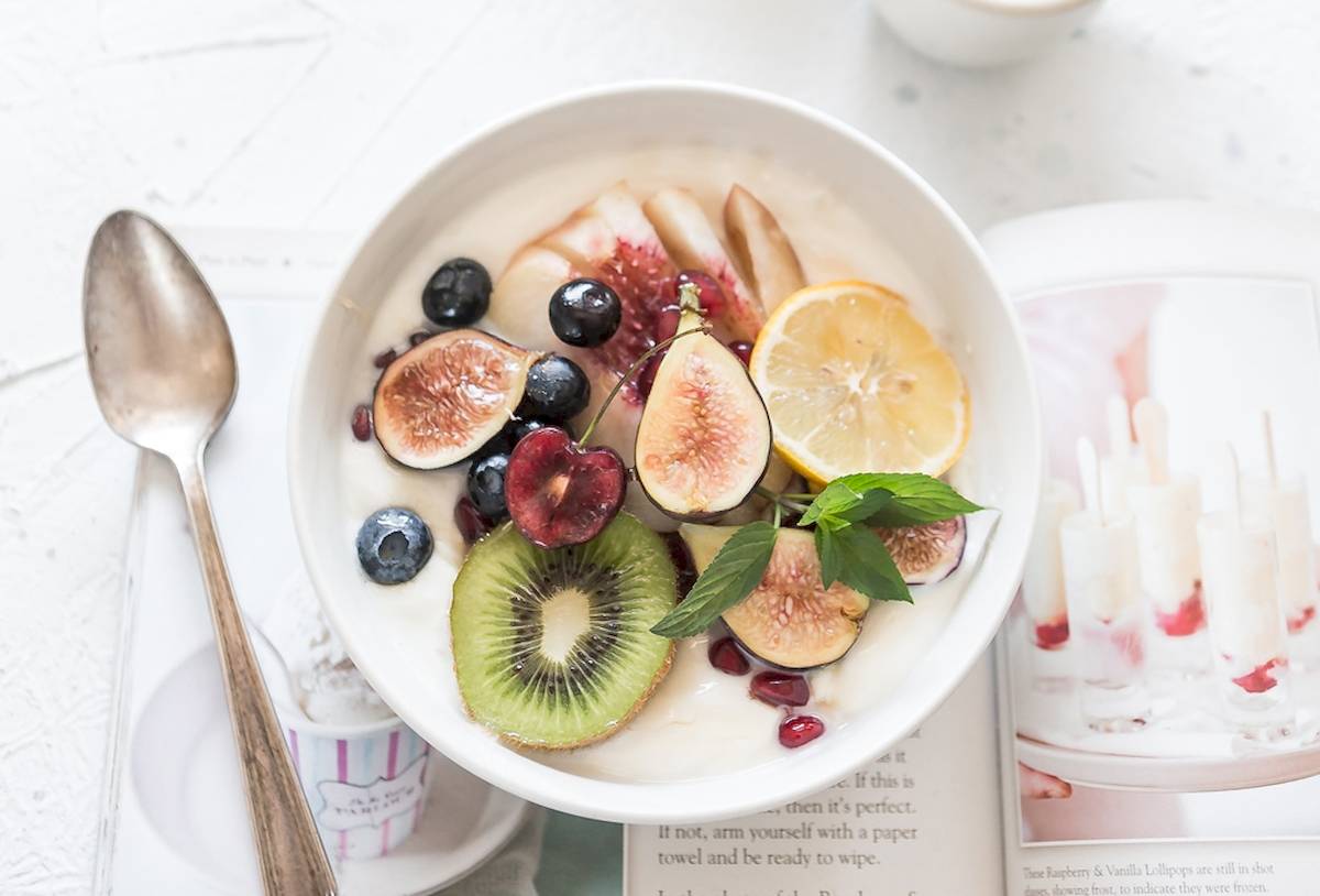 Ein weißer Schüssel mit Joghurt, gefüllt mit verschiedenen Früchten wie Feigen, Blaubeeren, Kiwis und Pomegranate, dazu ein Löffel und ein Buch.