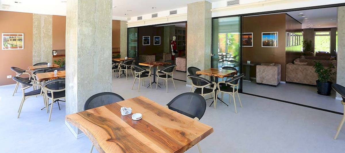 Innenbereich des Dyrrah Hotels mit mehreren Holztischen und Stühlen, großen Glastüren und einem Pflanzgefäß.