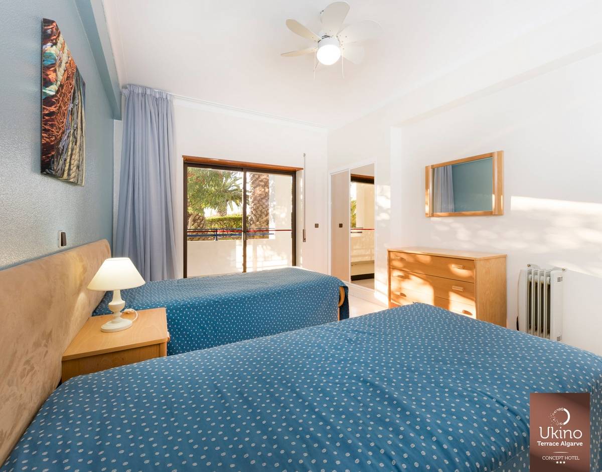 Innenansicht eines Doppelzimmers im Ukino Terrace Algarve Hotel mit zwei Betten, Nachttischen, Deckenventilator und Fenstern.