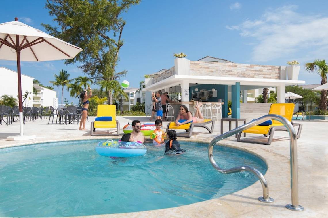 Familie, die im Swimmingpool des Emotions By Hodelpa Puerto Plata schwimmt, mit Blick auf einen Barbereich.