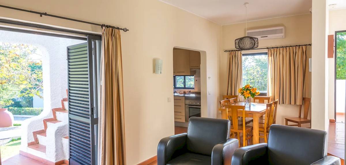 Wohnzimmer des Pedras da Rainha Resorts mit offenem Eingang, Tisch mit Blumen und Blick auf die Außenterrasse.