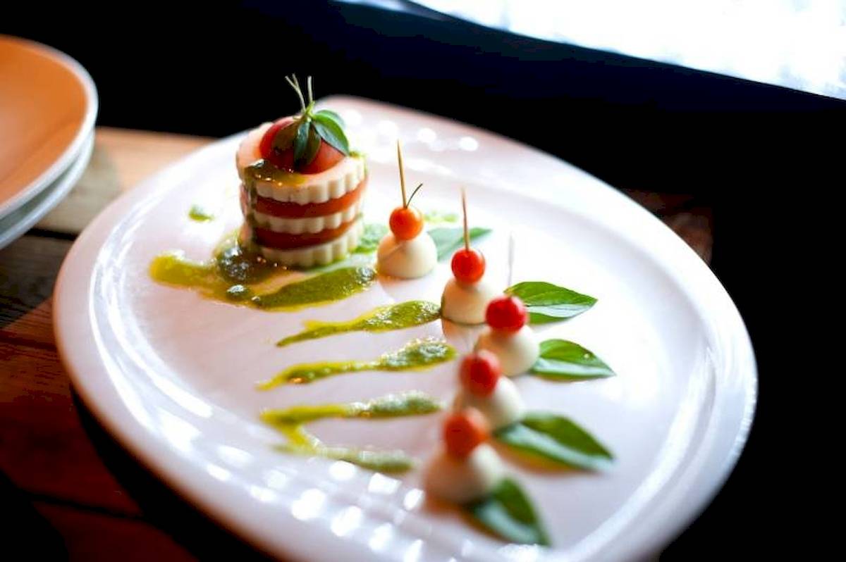 Ein elegantes Gericht auf einem weißen Teller, bestehend aus mehreren Schichten von Tomaten und Mozzarella, dekoriert mit roten Tomaten und grünen Blättern.