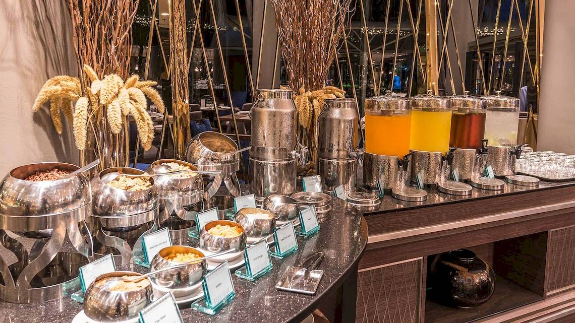 Buffetstation im Chatrium Hotel Riverside Bangkok mit verschiedenen Getränken und Speisen in silbernen Behältern.