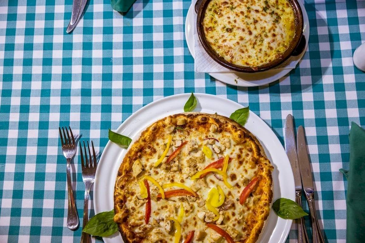 Eine Pizza auf einem Teller mit einem kleinen Topf auf einem blau-weiß karierten Tisch.