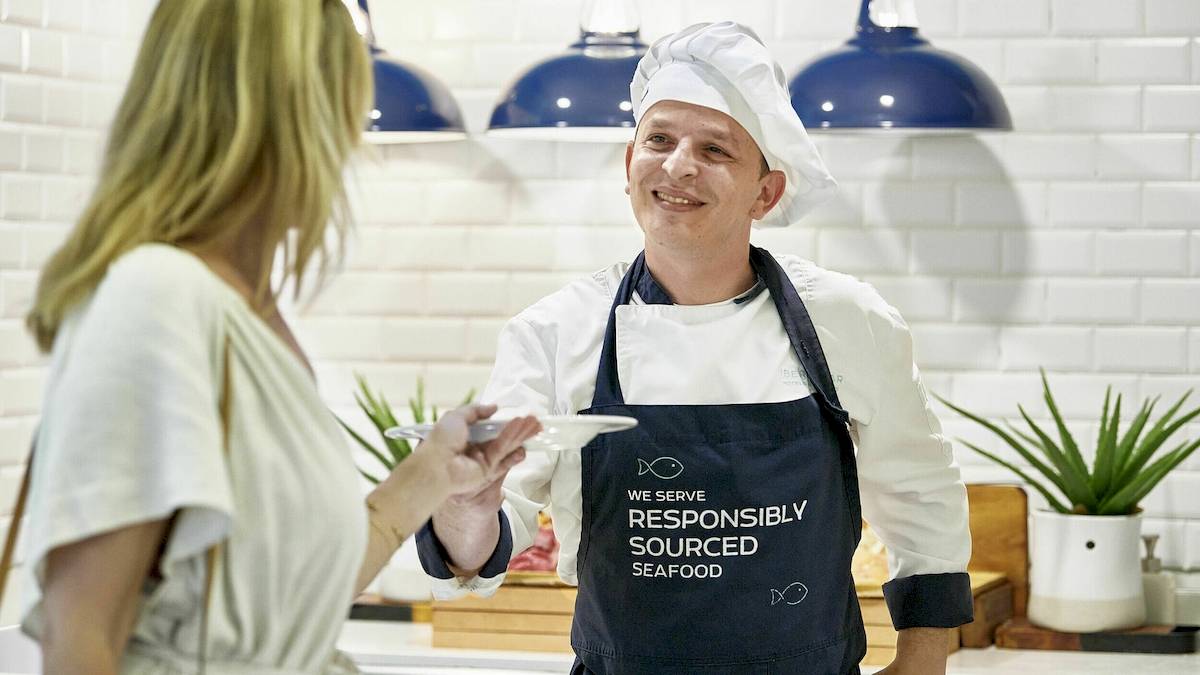 Koch mit Apron, das 'Wir servieren verantwortungsvoll beschafften Fisch' trägt, gibt einer Frau Essen im Iberostar Cristina Hotel.