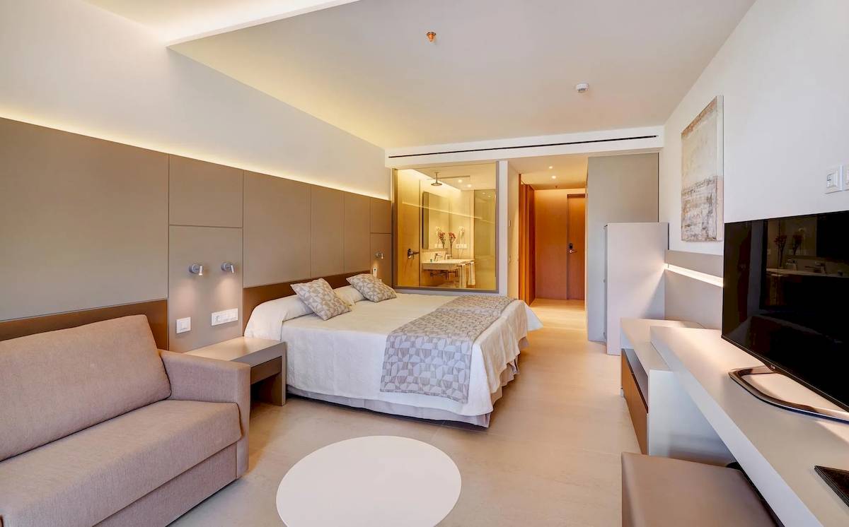 Innenansicht eines Hotelzimmers im Hipotels Gran Playa de Palma, mit Bett, Sofa, Fernseher und modernem Badezimmer.