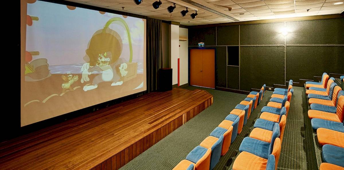 Kinossaal im Gloria Serenity Resort mit großen Leinwand und Reihen von orange und blauen Stühlen.