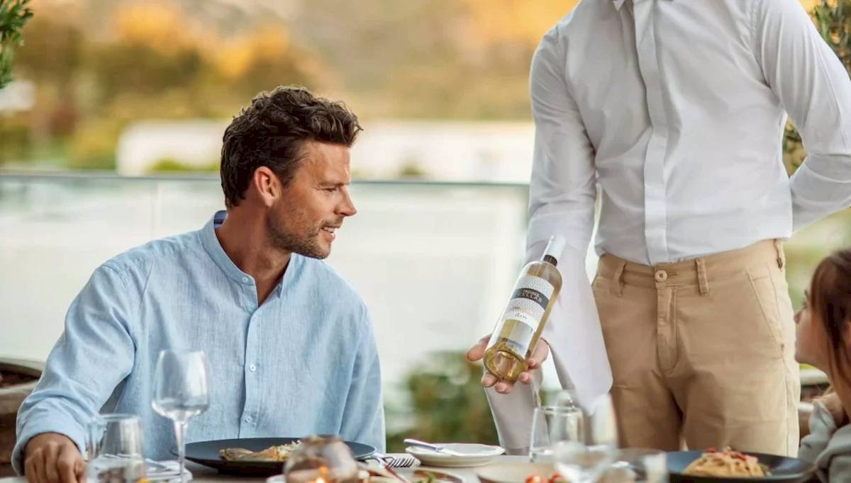 Ein Mann öffnet eine Flasche Wein an einem Tisch, während ein Kellner ein Glas einschenkt.