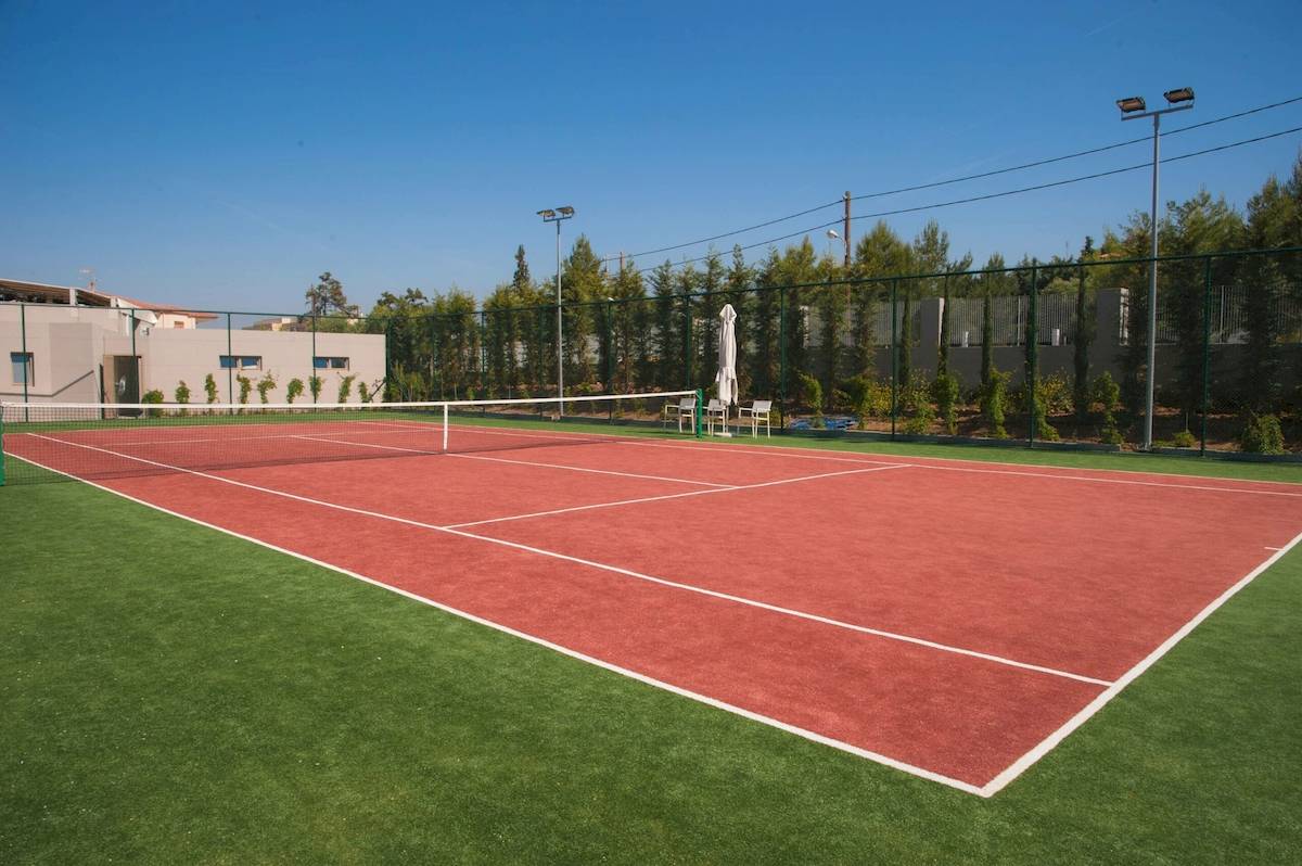 Tennisplatz im Ikos Dassia Hotel mit rotem Boden und grünem Rasen.