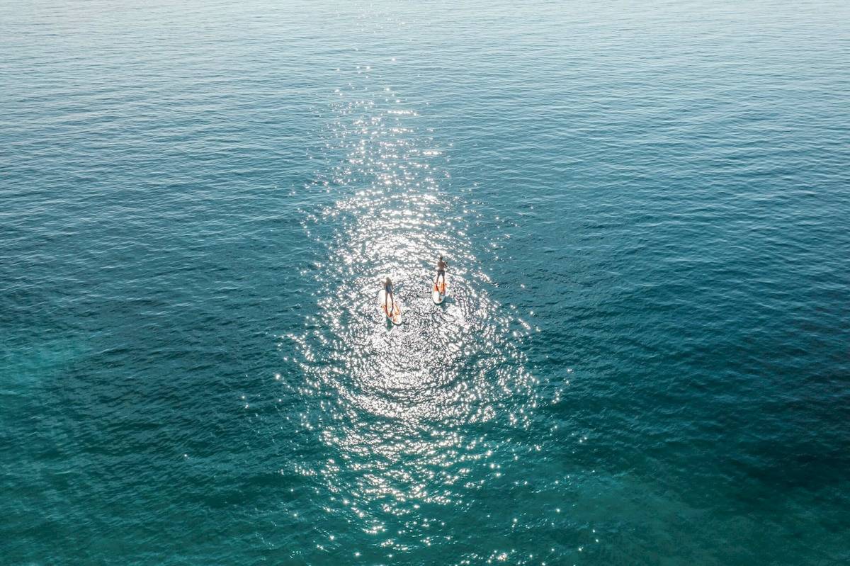Zwei Personen auf Surfbrettern im kristallklaren Wasser vor einer blauen Himmelslandschaft.
