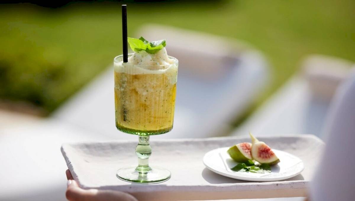 Ein Glas mit einem gelb-grünen Getränk und einem Strohhalm, serviert auf einem Tablett mit einer kleinen Platte mit Feigen.