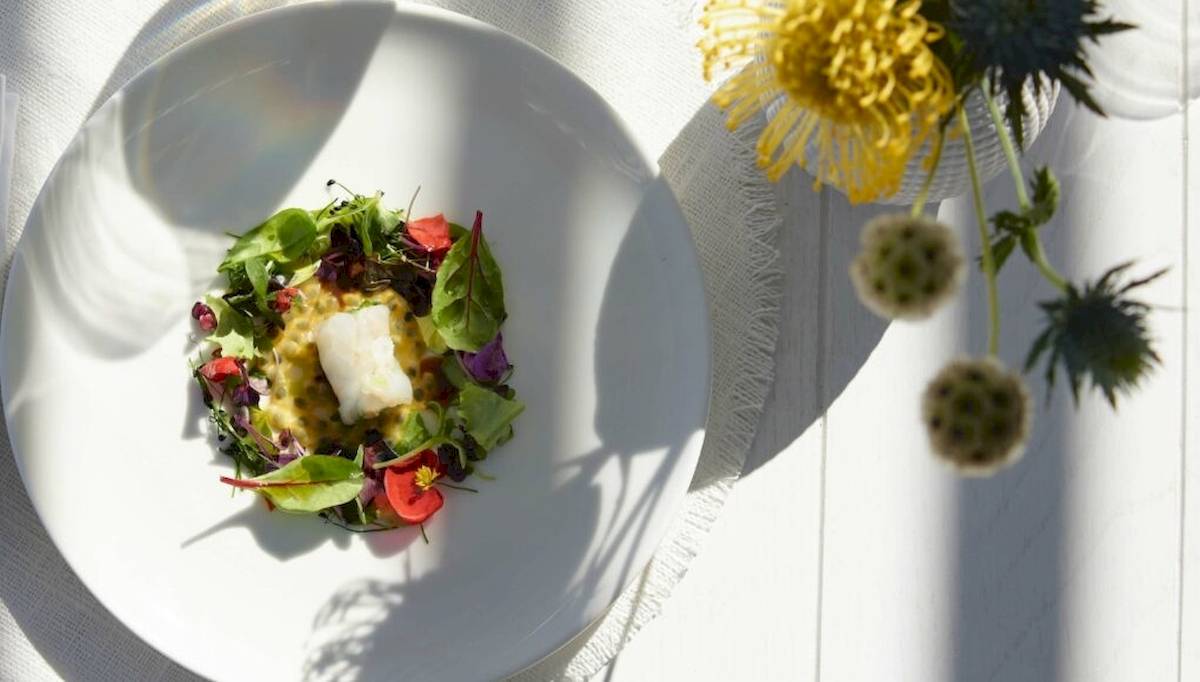 Ein Salat mit Fisch auf einem weißen Teller, umgeben von bunten Blüten auf einem Tisch.